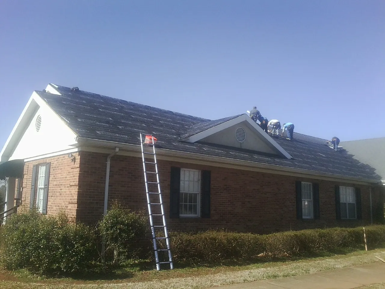 Crew tearing shingles off a brick ranch roof with a ladder set — Emergency Roof Repair work by our crew in Bostic