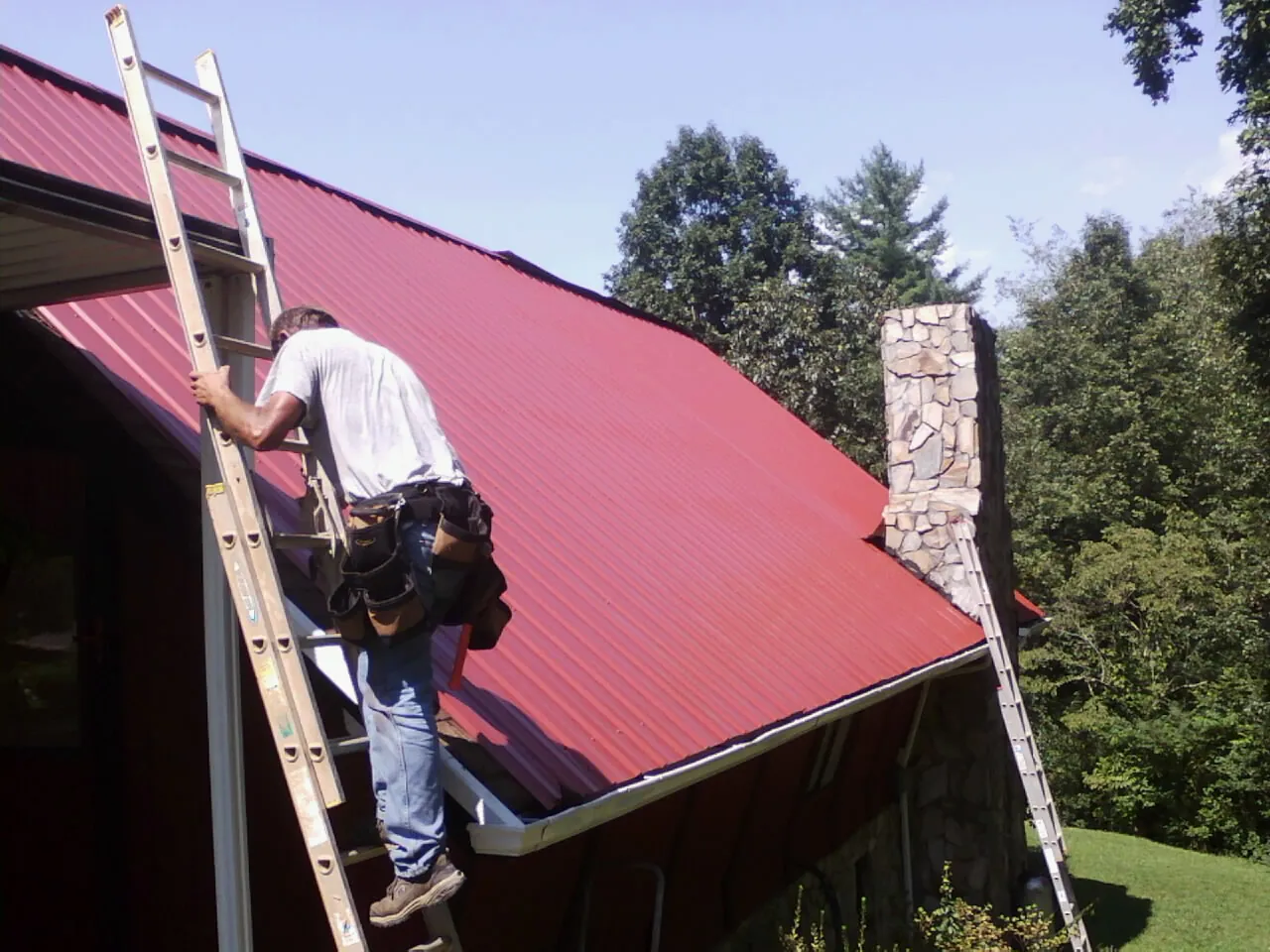 Roofer on a ladder beside a red metal roof and stone chimney — Soffit & Fascia Repair by Roofing Brothers in Bostic